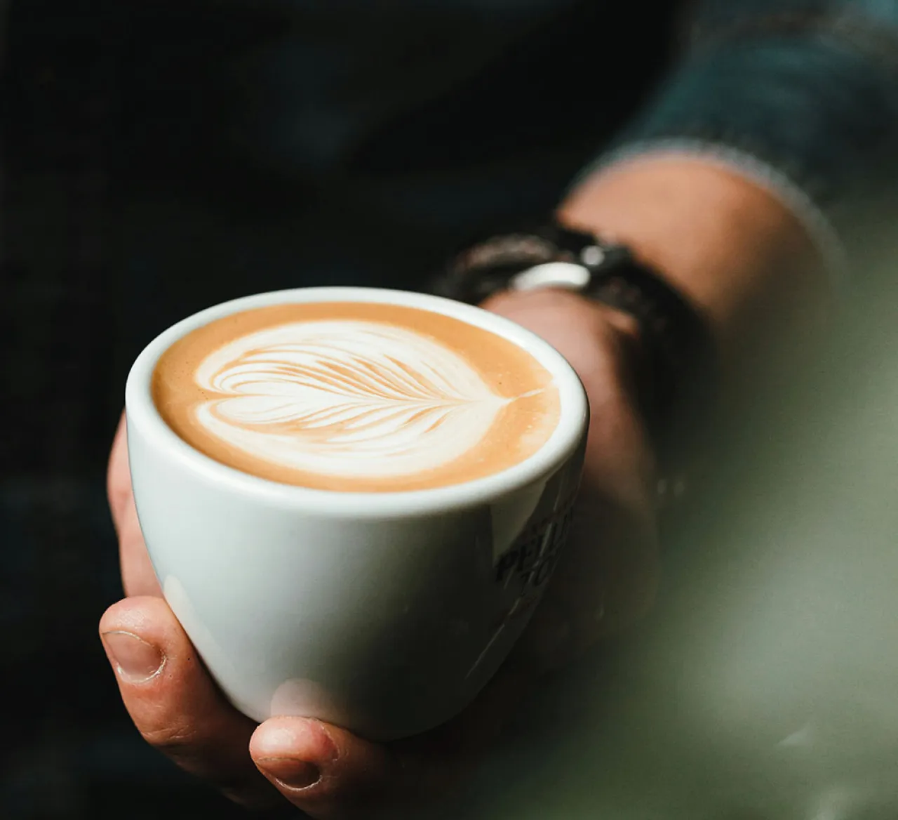 Hand hält eine Tasse Cappuccino mit kunstvoller Latte Art in Herzform. Warme Lichtstimmung, cremiger Milchschaum und detailreiche Nahaufnahme für echten Kaffeegenuss.